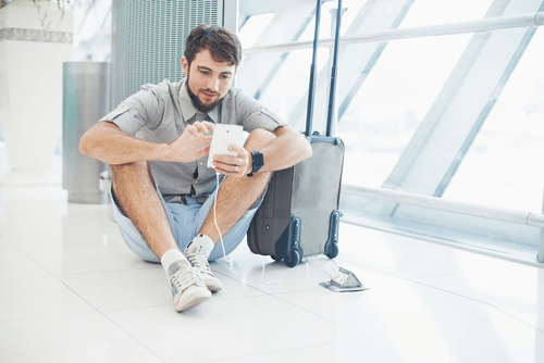 white man in casual clothing sitting on the floor holding a cellphone in his hand with his luggage by his side