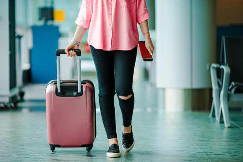 Woman in casual clothing at the airport with a pink suitcase in one hand and a cellphone in the other
